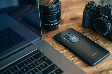 Laptop, phone and camers lens on a wooden table top.