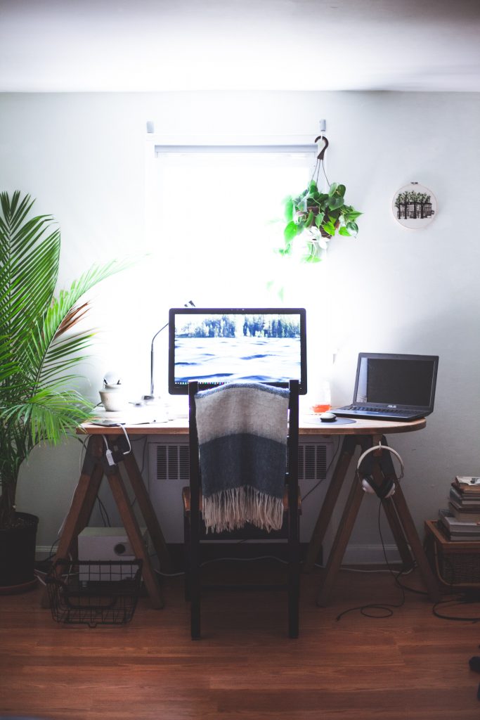 Home office desk with a monitor, laptop, and headphones by a bright window, flanked by houseplants