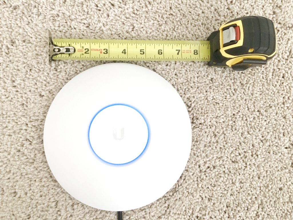 White Ubiquiti UniFi AP AC Pro access point on carpet beside a tape measure for scale