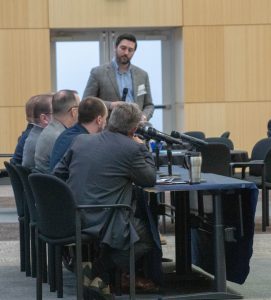 Executive panel moderator standing to address panelists seated at a table with microphones
