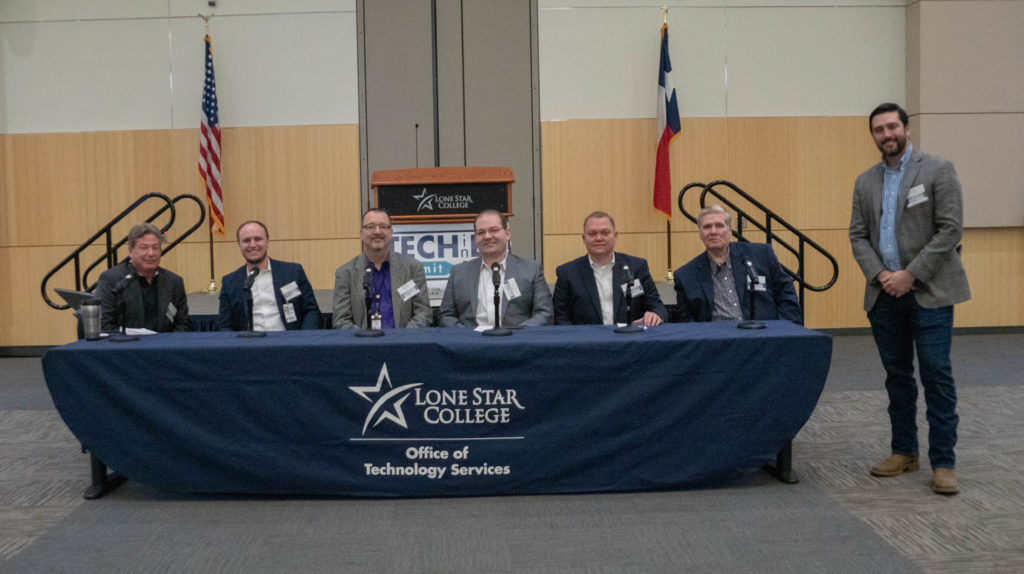 Six executive panelists seated at a Lone Star College Office of Technology Services table, with the moderator standing